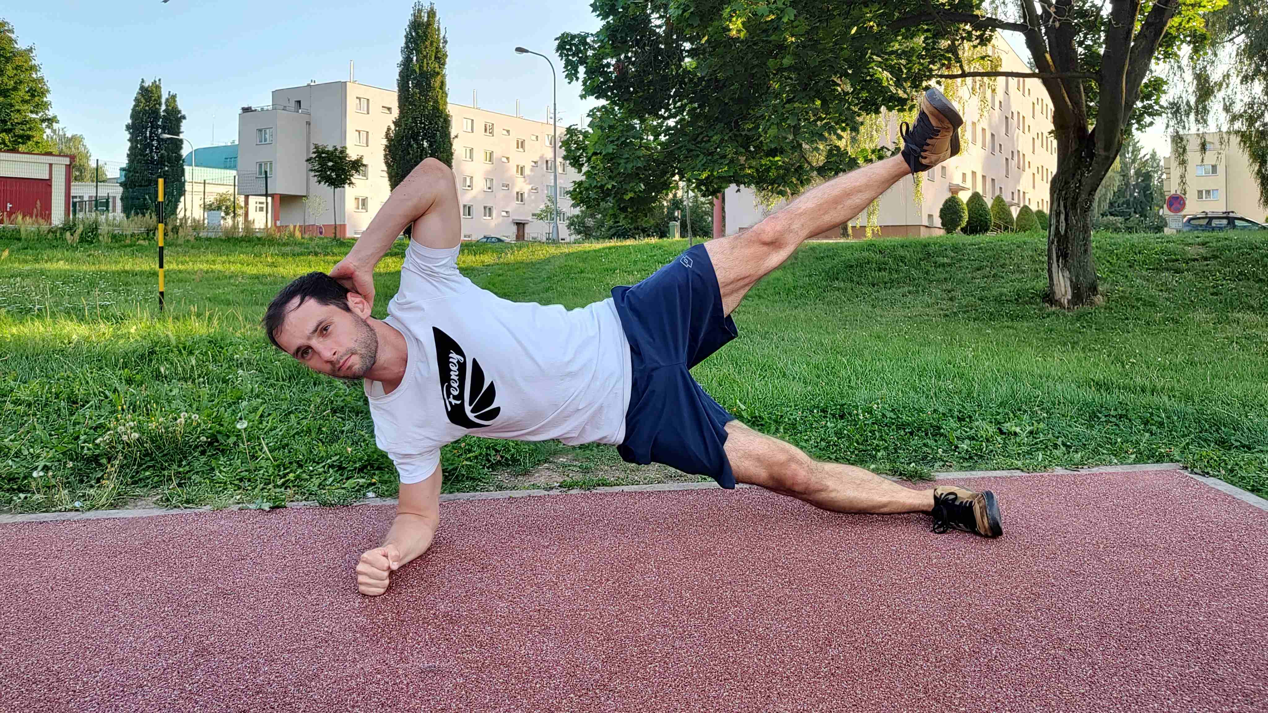 Muž v parku cvičí boční plank a při něm zvedá nohu do vzduchu.